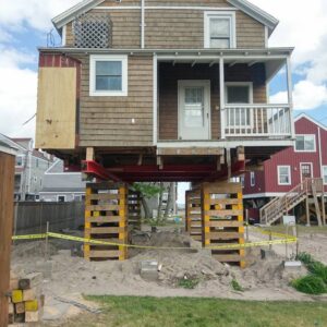 A house with wood shingle siding pulled up out of the ground and balancing on beams.